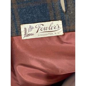 May include: A garment interior featuring a blue and brown plaid fabric lining and a white rectangular label. The label displays a thistle illustration and the text "Fowler's BINGHAMTON, N.Y." in black script and block lettering, set against a solid rust-colored fabric.
