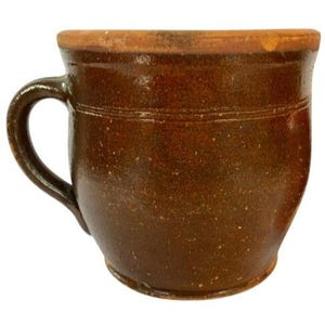 May include: A brown earthenware crock with a single handle. The crock has a rounded body and a slightly flared rim.