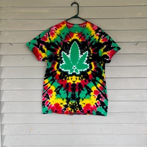 May include: A tie-dye t-shirt with a large green cannabis leaf graphic. The shirt features a vibrant pattern of red, yellow, green, and black colors. The t-shirt is hanging on a black hanger against a gray wooden background.