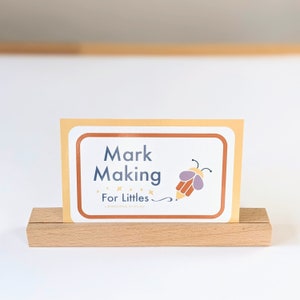 Peut inclure: Un présentoir en bois clair avec une carte affichant le texte "Mark Making For Littles" en bleu. La carte a une bordure jaune et une illustration d'abeille de dessin animé.