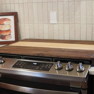 Puede incluir: Una cubierta rectangular de estufa de madera con una franja de madera clara en el centro. La cubierta se asienta sobre una estufa de acero inoxidable con perillas plateadas. Una foto enmarcada de donas y una cafetera están en el fondo.