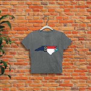 May include: A gray cropped t-shirt with a graphic of the state of North Carolina in the shape of the state flag. The flag is red, white, and blue with a yellow star in the blue triangle.