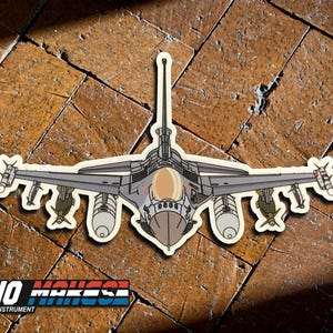 May include: A gray and white fighter jet sticker with a brown background. The jet is facing forward with its wings spread out. The sticker is on a tile floor.