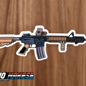 May include: A sticker of a black and tan AR-15 style rifle with a tan stock and a red dot sight. The sticker has a white outline and is on a wood background. The text "DEANO MAKES" is visible in the bottom left corner of the image.