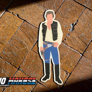 May include: A sticker featuring a cartoon illustration of Han Solo from Star Wars. He is wearing a brown vest, a white shirt, blue jeans, and brown boots. He is holding a blaster in his right hand. The sticker is on a brown tiled surface. The text "DEANO MAKES" is visible in the bottom left corner of the image.