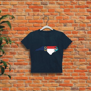 May include: A navy blue cropped t-shirt with a graphic of the state of North Carolina outlined in white and filled with the North Carolina state flag colors. The flag is red, white, and blue with a yellow star in the center.