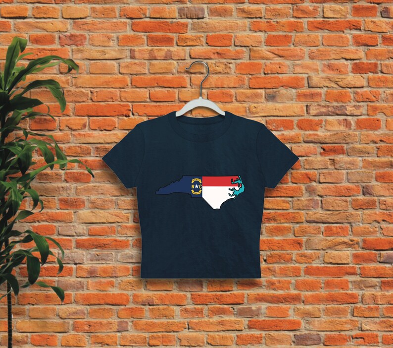 May include: A navy blue cropped t-shirt with a graphic of the state of North Carolina outlined in white and filled with the North Carolina state flag colors. The flag is red, white, and blue with a yellow star in the center.
