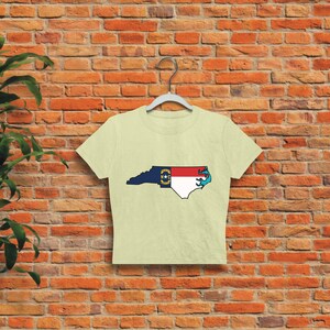 May include: A cream-colored cropped t-shirt with a graphic of the state of North Carolina outlined in blue and filled with the North Carolina state flag.