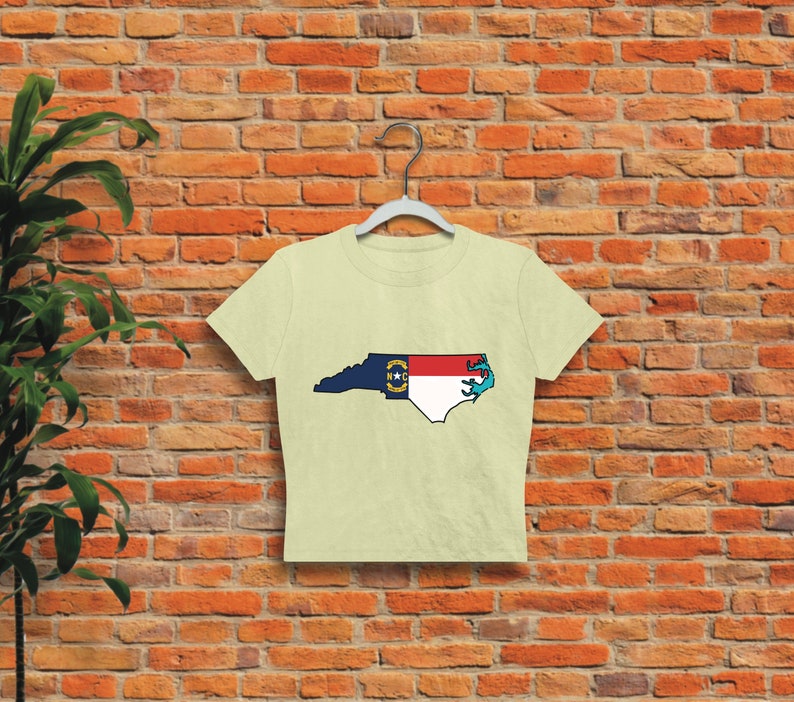 May include: A cream-colored cropped t-shirt with a graphic of the state of North Carolina outlined in blue and filled with the North Carolina state flag.