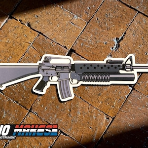 May include: A black and gray cartoon illustration of a rifle with a grenade launcher attached. The rifle is on a brown tiled surface. The image includes the text "DEANO MAKES" in blue, white, and red.