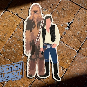 May include: A sticker featuring a brown and white Wookiee character from Star Wars standing next to a human character wearing a brown vest and blue jeans. The sticker is on a brown tiled surface.