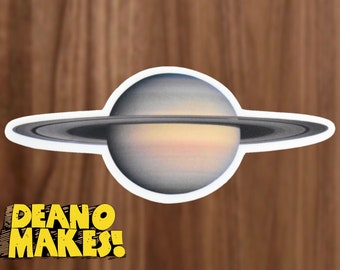 May include: A sticker of Saturn, a gas giant planet with a ring system, on a woodgrain background. The planet is depicted in shades of gray and white, with a faint yellow band around the equator. The rings are dark gray and white.