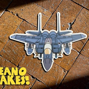 May include: A sticker featuring a blue and gray fighter jet with a white border. The sticker has a cartoon style and is set against a brown, textured background. The text "DEANO MAKES!" is in yellow.