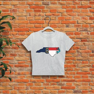 May include: A grey cropped t-shirt with a graphic of the state of North Carolina outlined in black and filled with the North Carolina state flag colors. The flag is red, white, and blue with a yellow star in the center.