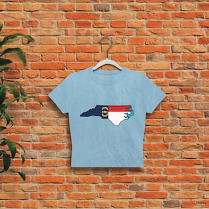 May include: A light blue cropped t-shirt with a graphic of the state of North Carolina, featuring the state flag. The outline of the state is filled with the colors of the North Carolina flag.