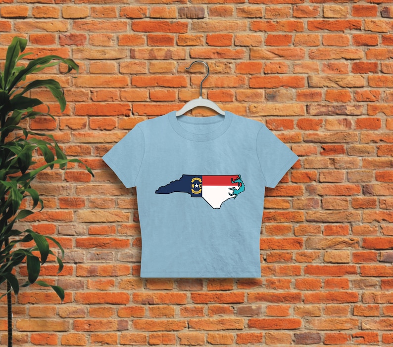May include: A light blue cropped t-shirt with a graphic of the state of North Carolina, featuring the state flag. The outline of the state is filled with the colors of the North Carolina flag.