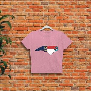 May include: A pink cropped t-shirt with a graphic of the state of North Carolina, featuring the state flag. The outline of the state is filled with the colors of the North Carolina flag.