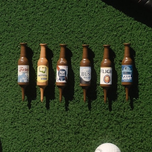 Pu&ograve; includere: Sei tee da golf in plastica marrone a forma di bottiglie di birra in miniatura, ognuna con un'etichetta unica con scritte tra cui Forest Light, Twisted Tea, Par Beer, Bush Right, Flight e Putty Light, disposte in fila su una superficie verde.