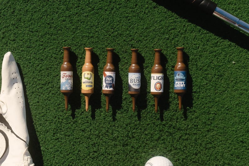 Pu&ograve; includere: Sei tee da golf in plastica marrone a forma di bottiglie di birra in miniatura, ognuna con un'etichetta unica con scritte tra cui Forest Light, Twisted Tea, Par Beer, Bush Right, Flight e Putty Light, disposte in fila su una superficie verde.