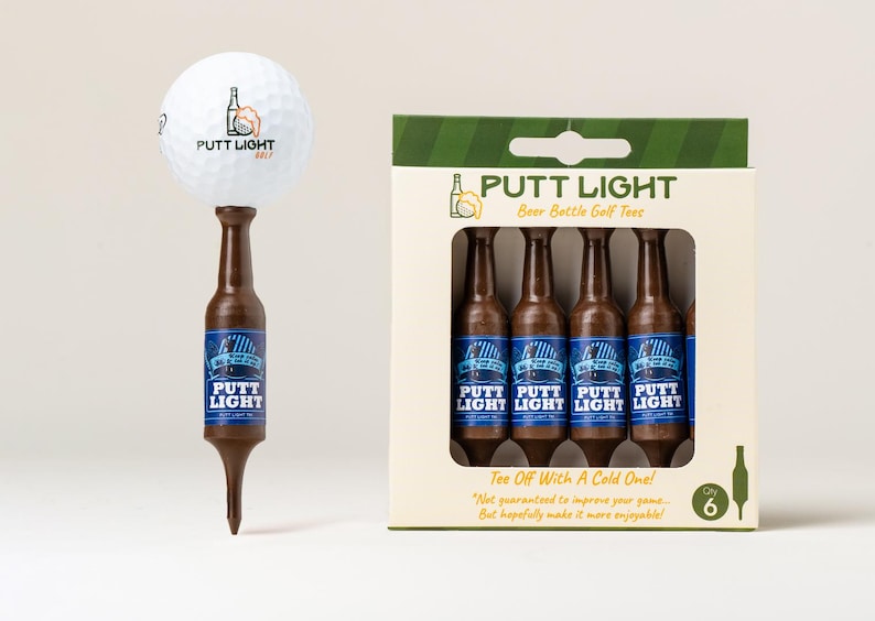 Pu&ograve; includere: Una pallina da golf bianca con il logo di una bottiglia di birra poggia su un tee da golf marrone a forma di bottiglia. Accanto, una scatola di cartone contiene sei tee da golf marroni identici a forma di bottiglia, etichettati Putt Light.