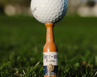 Può includere: Una palla da golf bianca è posizionata su un tee da golf marrone a forma di bottiglia di birra. La bottiglia è etichettata "Fore-on Extra" e "Putter-Up Golf Co."