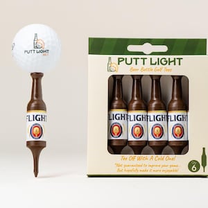 Pu&ograve; includere: Una pallina da golf bianca appoggiata su un tee da golf marrone a forma di bottiglia di birra, accanto a una scatola di cartone contenente quattro tee identici. La confezione riporta il testo Putt Light Beer Bottle Golf Tees e Tee Off With A Cold One.