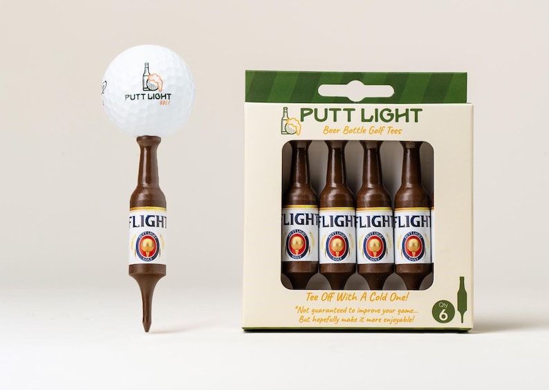 Pu&ograve; includere: Una pallina da golf bianca appoggiata su un tee da golf marrone a forma di bottiglia di birra, accanto a una scatola di cartone contenente quattro tee identici. La confezione riporta il testo Putt Light Beer Bottle Golf Tees e Tee Off With A Cold One.