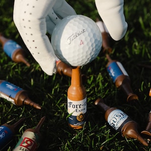 Pu&ograve; includere: Una pallina da golf bianca Titleist 4 in equilibrio su un tee da golf a forma di bottiglia di birra marrone con un'etichetta che riporta la scritta Fore-O Extra. Altri tee simili sono sparsi sull'erba verde.