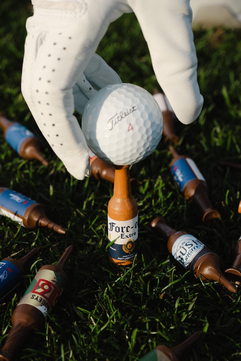Pu&ograve; includere: Una pallina da golf bianca Titleist 4 in equilibrio su un tee da golf a forma di bottiglia di birra marrone con un'etichetta che riporta la scritta Fore-O Extra. Altri tee simili sono sparsi sull'erba verde.