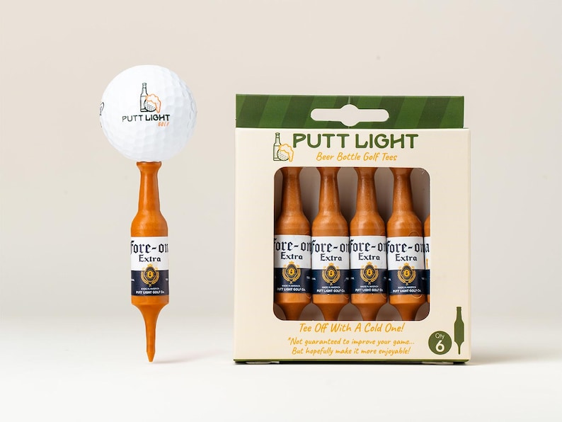 Pu&ograve; includere: Una pallina da golf bianca appoggiata su un tee da golf marrone a forma di bottiglia di birra, accanto a una scatola di cartone contenente sei tee identici. La confezione riporta il testo PUTT LIGHT, Beer Bottle Golf Tees, Fore-on Extra e Tee Off With A Cold One.