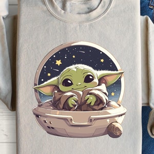May include: A light beige sweatshirt with a graphic print of a green baby Yoda character in a spaceship against a dark blue background with stars.