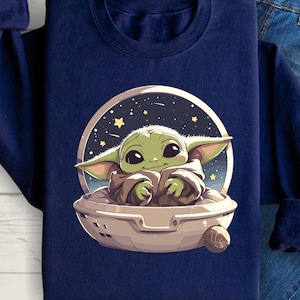 Puede incluir: Una sudadera azul marino con un gr&aacute;fico de un personaje alien&iacute;gena verde sentado en una nave espacial sobre un fondo de cielo nocturno estrellado.