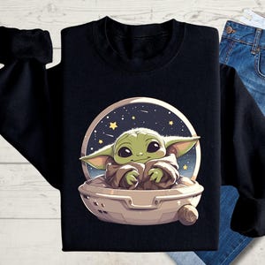 Puede incluir: Sudadera negra con un personaje alien&iacute;gena verde en una nave espacial, sobre un fondo de cielo nocturno estrellado.