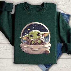 Puede incluir: Una sudadera verde oscuro con un gr&aacute;fico de un personaje alien&iacute;gena verde en una nave espacial contra un fondo de cielo nocturno estrellado.