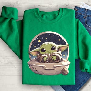 Puede incluir: Sudadera verde con una imagen de dibujos animados de un personaje alien&iacute;gena verde en una nave espacial. El extraterrestre sonr&iacute;e y tiene ojos grandes. La nave espacial est&aacute; rodeada de estrellas.