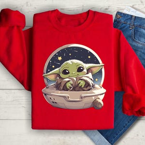 Puede incluir: Sudadera roja con un gr&aacute;fico de un personaje de beb&eacute; alien&iacute;gena verde en una nave espacial sobre un fondo estrellado.