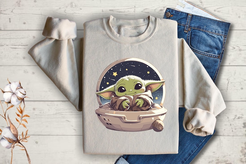 May include: A light beige sweatshirt with a graphic print of a green baby Yoda character in a spaceship against a dark blue background with stars.