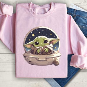 Puede incluir: Sudadera rosa con un gr&aacute;fico de un personaje alien&iacute;gena verde en una nave espacial sobre un fondo estrellado.