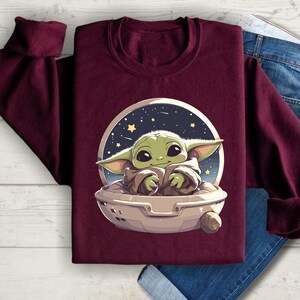 Puede incluir: Sudadera color burdeos con un gr&aacute;fico de un personaje alien&iacute;gena verde en una nave espacial. El personaje est&aacute; sentado en la nave espacial con los brazos cruzados y tiene una expresi&oacute;n linda en su rostro.