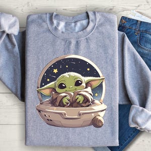 Puede incluir: Una sudadera gris con un estampado gr&aacute;fico de un personaje alien&iacute;gena verde con ojos grandes, sentado en una nave espacial sobre un fondo de cielo nocturno estrellado.