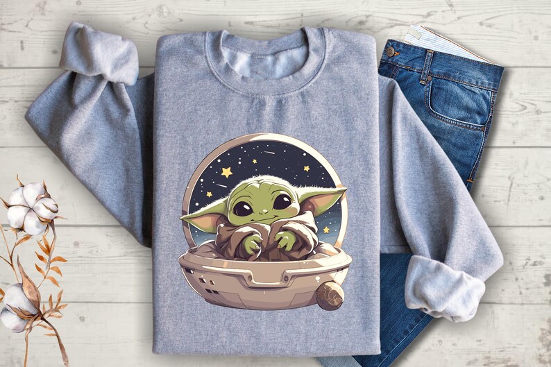 Puede incluir: Una sudadera gris con un estampado gr&aacute;fico de un personaje alien&iacute;gena verde con ojos grandes, sentado en una nave espacial sobre un fondo de cielo nocturno estrellado.