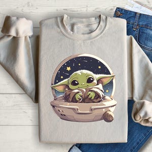 Puede incluir: Una sudadera beige con un gr&aacute;fico de un personaje alien&iacute;gena verde en una nave espacial sobre un fondo estrellado azul oscuro.