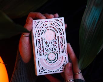 May include: A rectangular deck of playing cards featuring an intricate white laser-cut paper sleeve with arched architectural patterns and delicate pink accents. The design includes a circular emblem at the center and geometric filigree details.