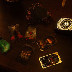 May include: A deck of tarot cards with ornate, dark imagery, scattered on a wooden surface. The cards feature illustrations with red, green, and gold accents. A matching box with a glowing gold design sits nearby, suggesting a mystical theme.