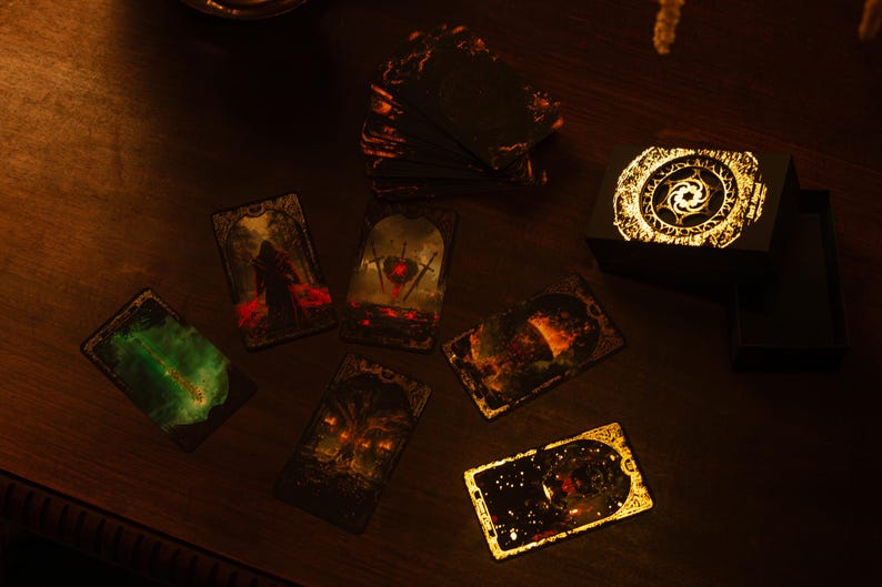 May include: A deck of tarot cards with ornate, dark imagery, scattered on a wooden surface. The cards feature illustrations with red, green, and gold accents. A matching box with a glowing gold design sits nearby, suggesting a mystical theme.