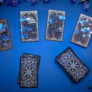 May include: A deck of tarot cards featuring dark, ornate borders and illustrations of blue lilies and metallic armour. Some cards are displayed face-up showing detailed artwork, while others are face-down revealing a circular, intricate white pattern.