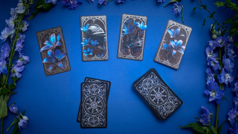 May include: A deck of tarot cards featuring dark, ornate borders and illustrations of blue lilies and metallic armour. Some cards are displayed face-up showing detailed artwork, while others are face-down revealing a circular, intricate white pattern.