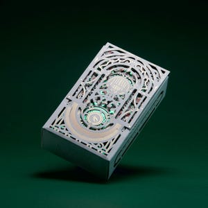May include: A rectangular playing card deck box featuring an intricate, laser-cut light grey outer sleeve with ornate filigree patterns. The design reveals a metallic green and gold inner layer with the text Eternal Garden printed on the front.