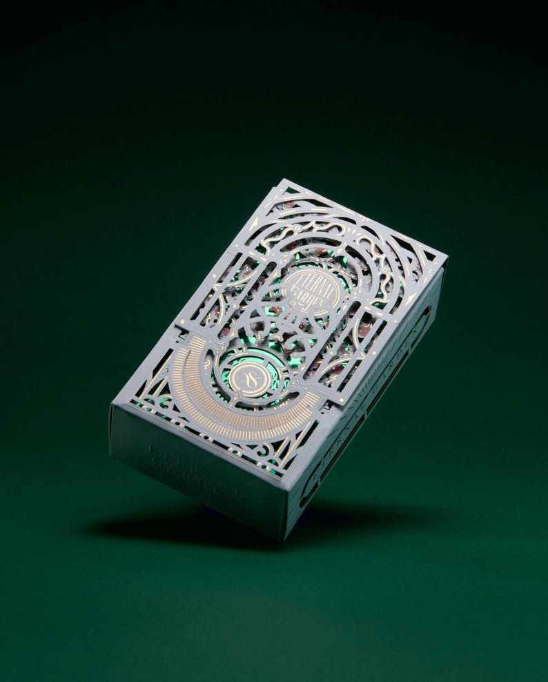 May include: A rectangular playing card deck box featuring an intricate, laser-cut light grey outer sleeve with ornate filigree patterns. The design reveals a metallic green and gold inner layer with the text Eternal Garden printed on the front.