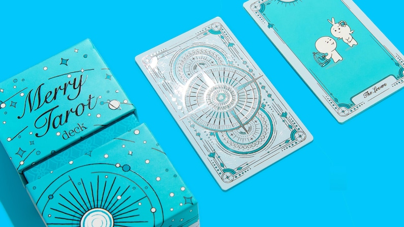 Może przedstawiać: Turkusowe pudełko i karty do tarota są wyświetlane na jasnoniebieskim tle. Na pudełku widnieje napis "Merry Tarot deck" w czcionce skryptowej. Karty zawierają ilustracje i wz&oacute;r kompasu. Jedna karta jest oznaczona jako "The Lovers".